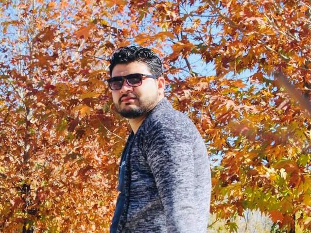 Ahmad looks at the camera while wearing sunglasses and standing in front of fall foliage.