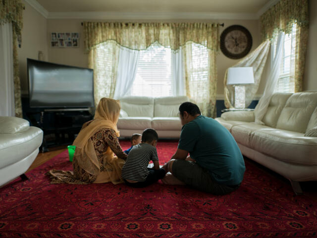 With their back to the camera, a husband and wife sit on their living room floor with their two children. The room also has a tv and white sofa.