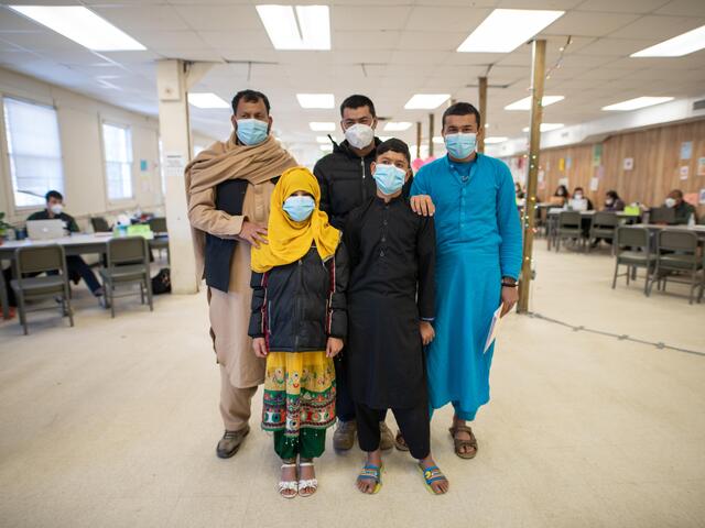 An Afgha family, all wearing masks, in a large room. There a three men, one boy and one girl.