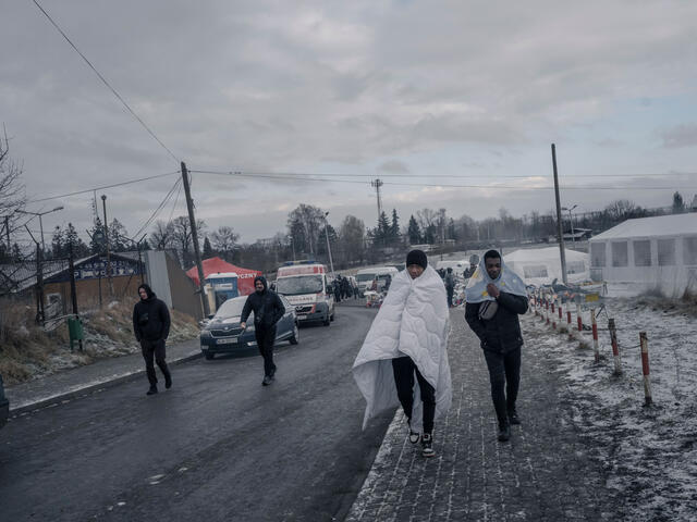 Two refugee men walk down the street with blankets over the shoulders.