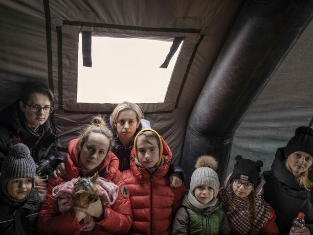 Women and children in winter clothes stand in a tent and look at the camera.