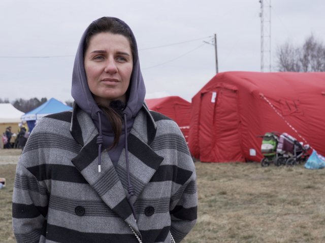 A woman stands in a reception area for refugees looking at the camera. There are tents in the background.