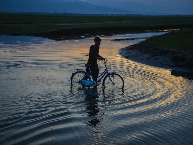 The silhouette of a man holding a bike in a shallow body of water in the evening.