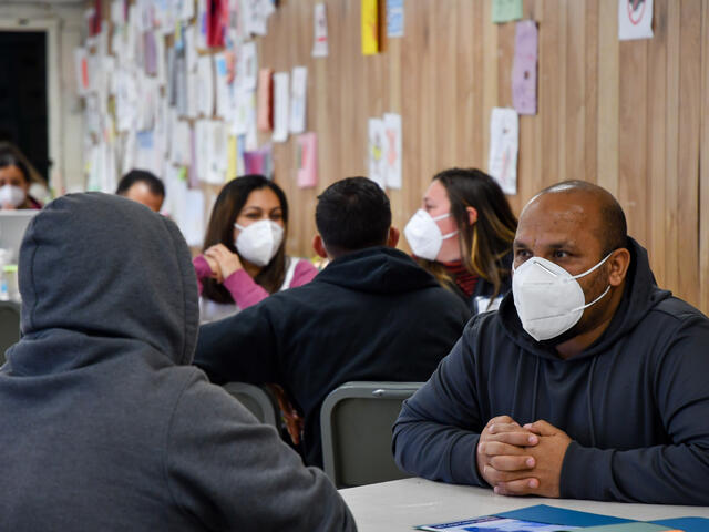 Wearing a mask, an IRC staff member sits at a table and talks to a client, whose back is to the camera.