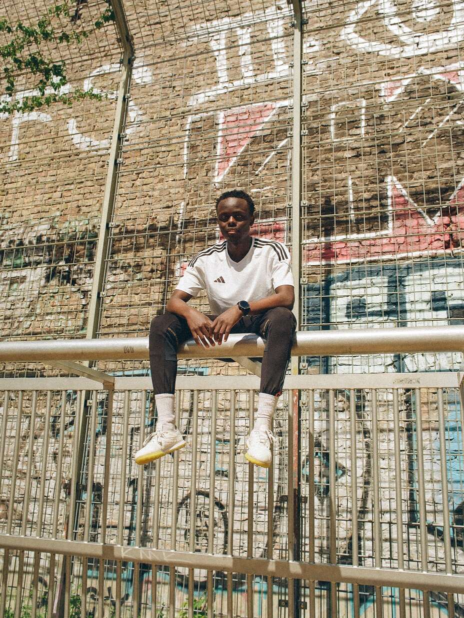 Ein Mann in einem weißen Hemd und dunklen Hosen sitzt auf einem Zaun vor einer mit Graffiti bedeckten Wand.