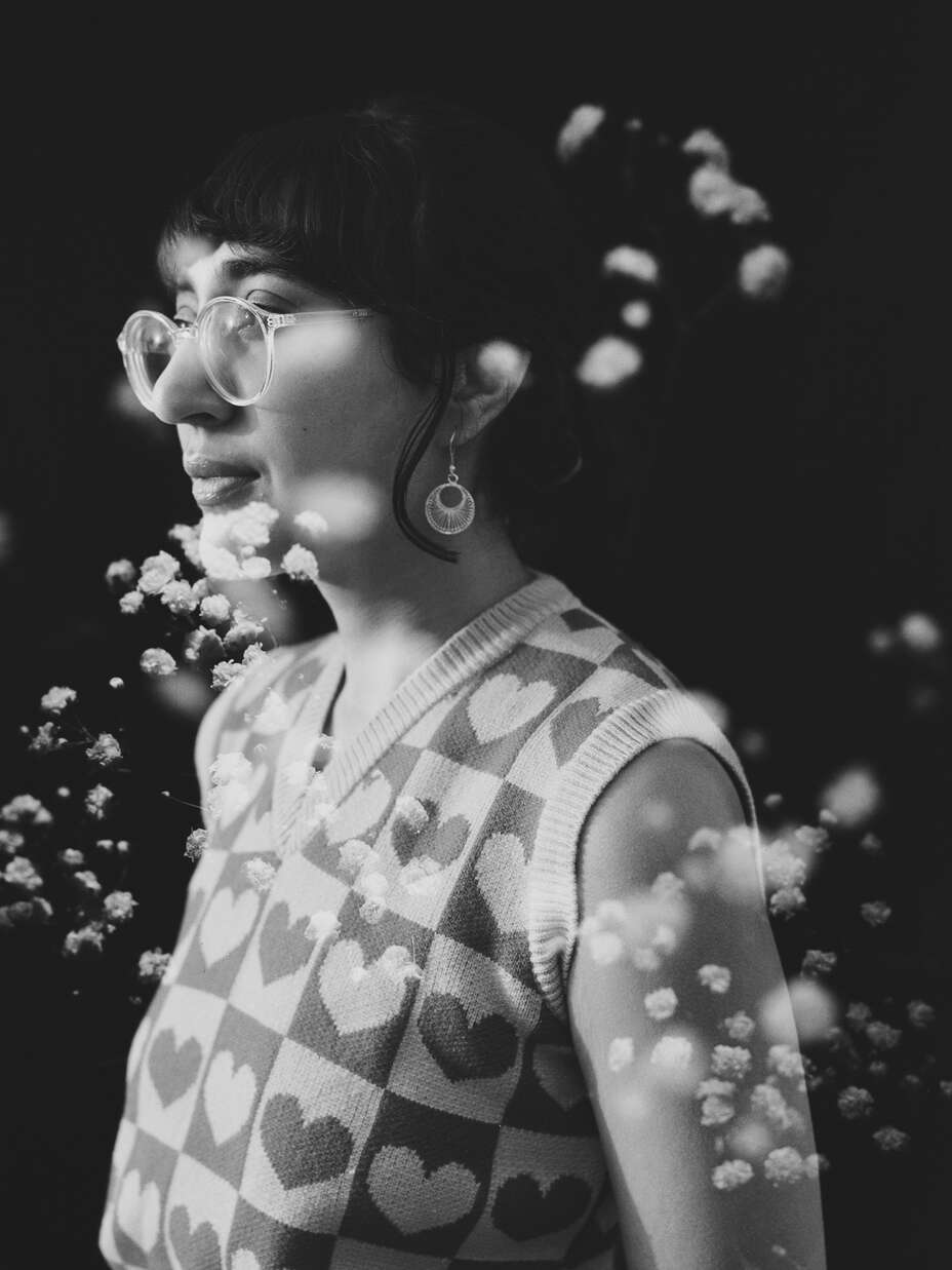Eine schwarz-weiße Aufnahme einer Person, die in einem ärmellosen Oberteil steht und von schwebenden Blütenblättern umgeben ist.
