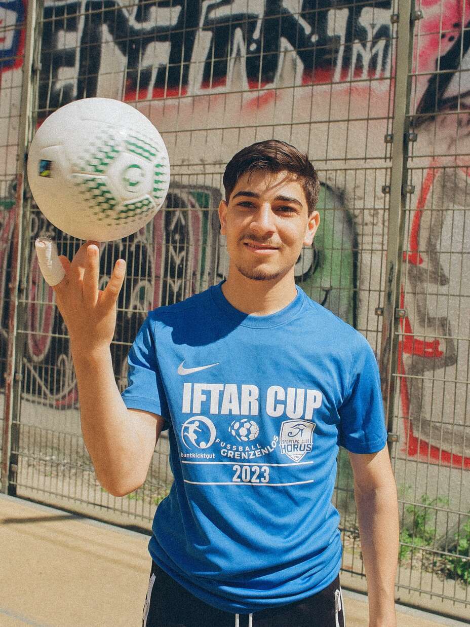 Ein junger Mann in einem blauen „Iftar Cup“-T-Shirt balanciert einen Fußball auf seinem Finger vor einer Wand mit Graffiti.