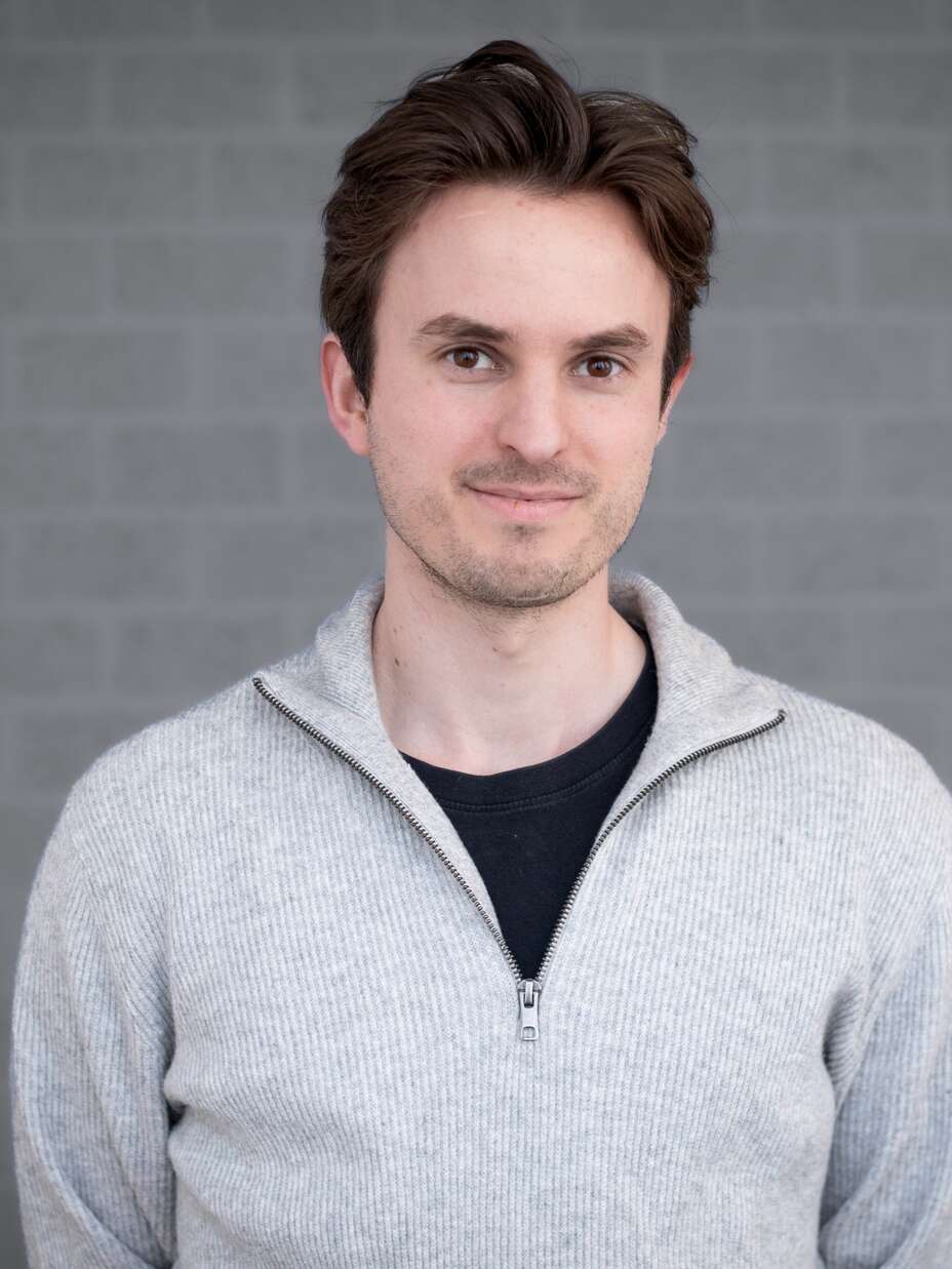 Ein Mann mit braunen Haaren steht vor einer grauen Backsteinmauer und schaut lächelnd in die Kamera.