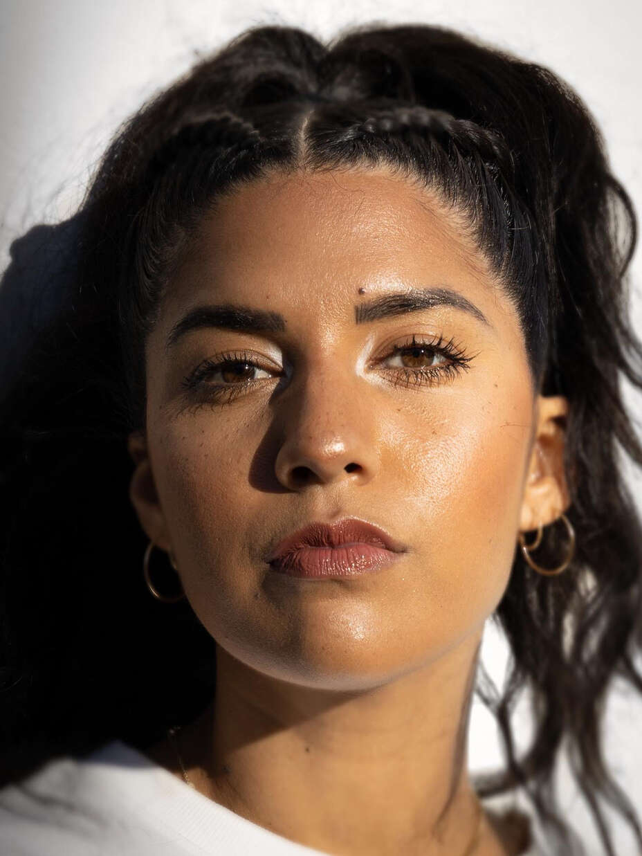 Eine Frau mit dunklem Haar und ernstem Gesichtsausdruck schaut direkt in die Kamera, beleuchtet von hellem Licht von der linken Seite.