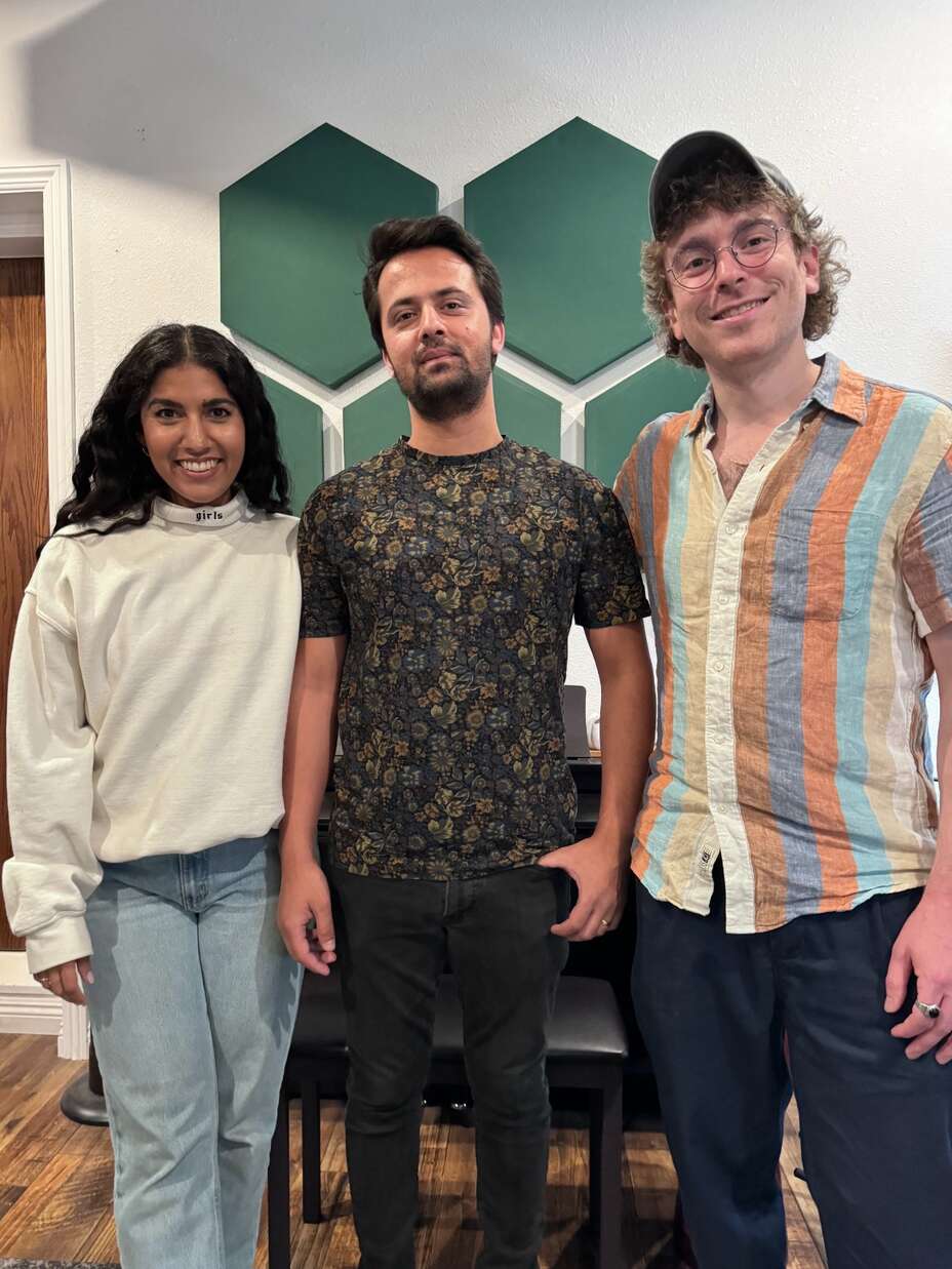 Drei Personen lächeln in die Kamera, zwei Männer und eine Frau, die vor einer grün-weißen Wabenstruktur stehen.