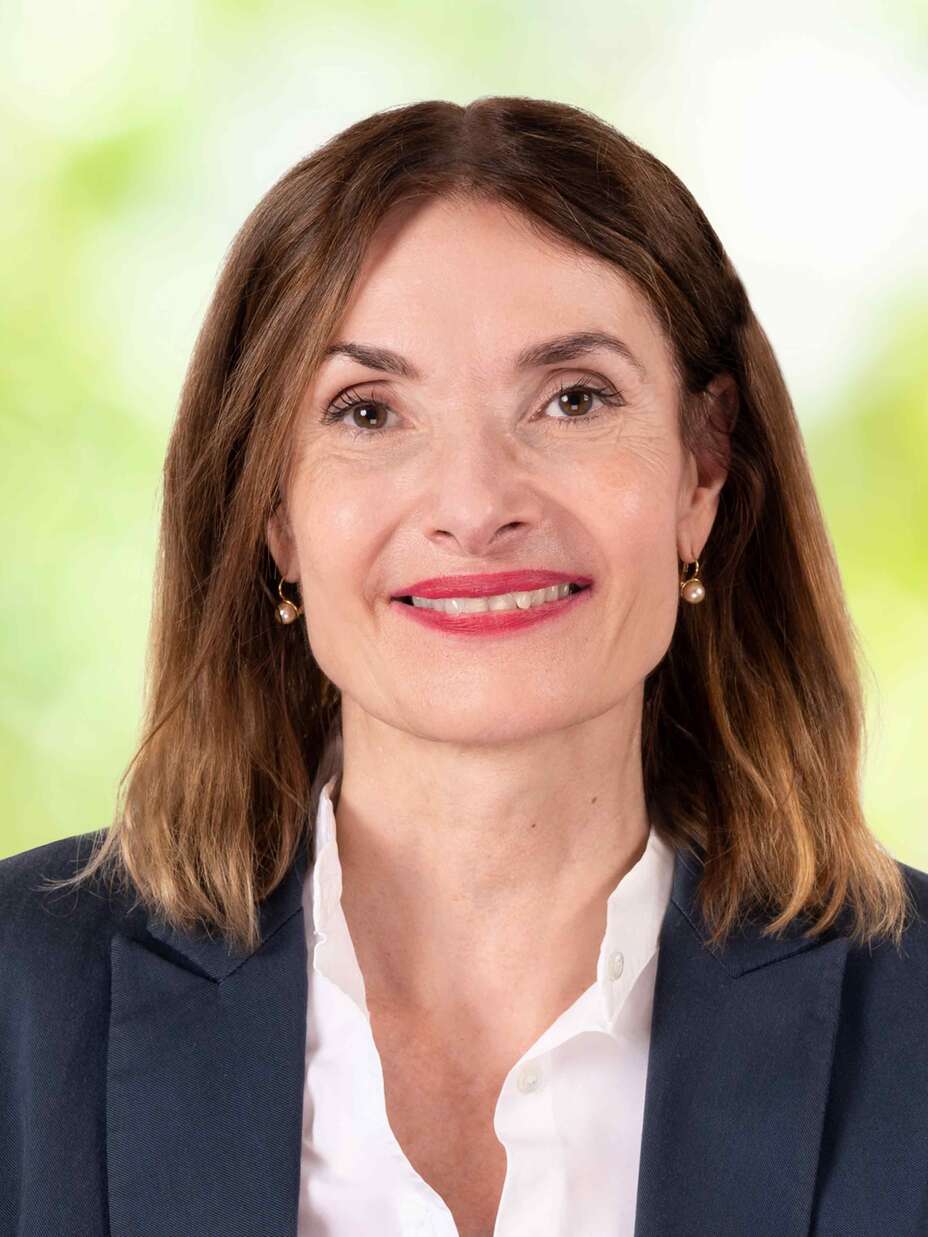 Eine Frau in einem Blazer lächelt vor einem verschwommenen, grünen Hintergrund in die Kamera.