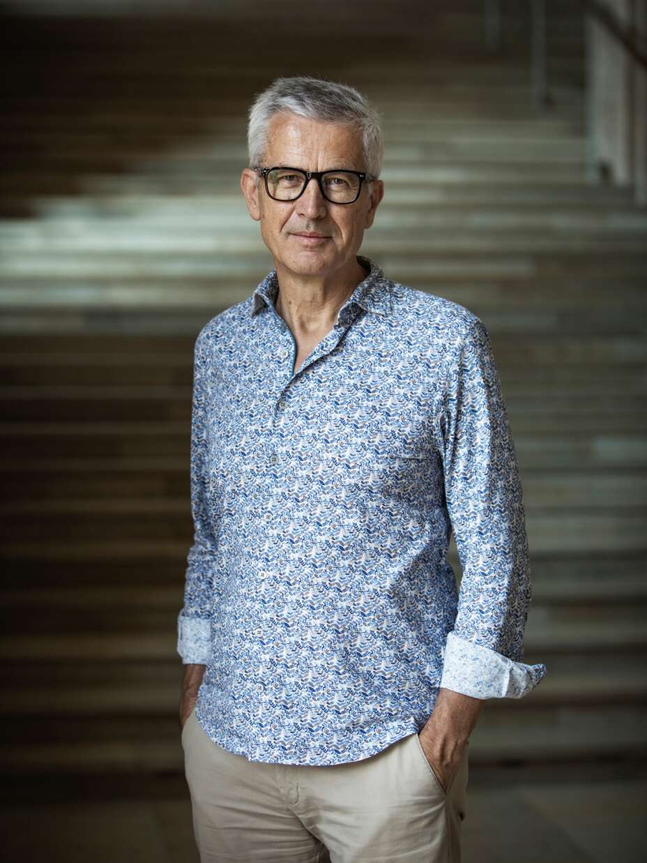 Ein Mann mit Brille steht entspannt vor einer beleuchteten Treppe, trägt ein hellblaues Hemd und eine beige Hose.