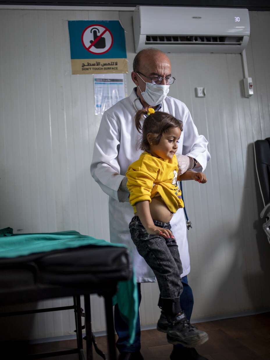 Ein Arzt in Schutzkleidung hält ein kleines Kind in gelber Kleidung in einer medizinischen Einrichtung, während er es vermutlich untersucht.