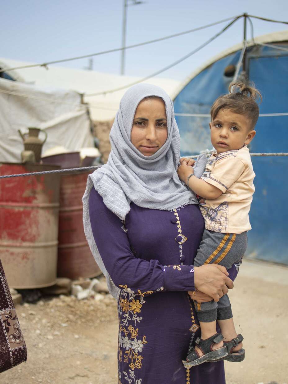 Eine Frau in einem blauen Kopftuch hält ein kleines Kind in einer Notunterkunft für Geflüchtete, umgeben von Zelten und Decken, die als Sichtschutz aufgehängt sind.