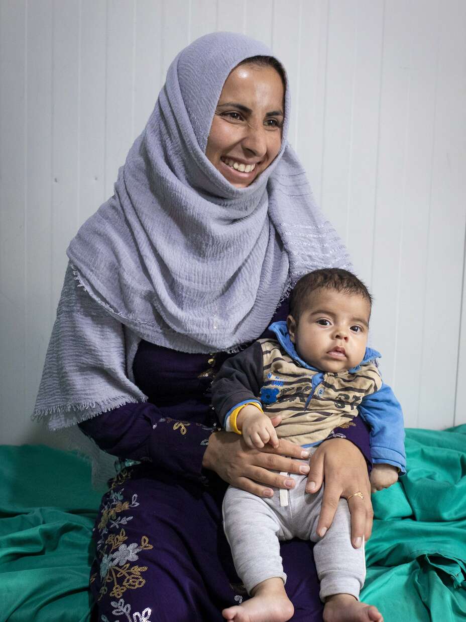 Eine lächelnde Frau in einem grauen Kopftuch hält ein Baby auf ihrem Schoß