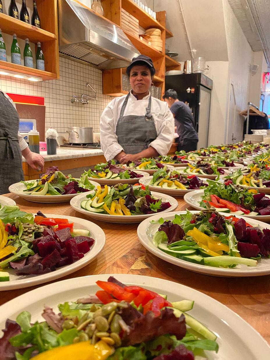 Wearing an apron, Chef Shanthini stands behind a table in a kitchen with many plates of a salad.