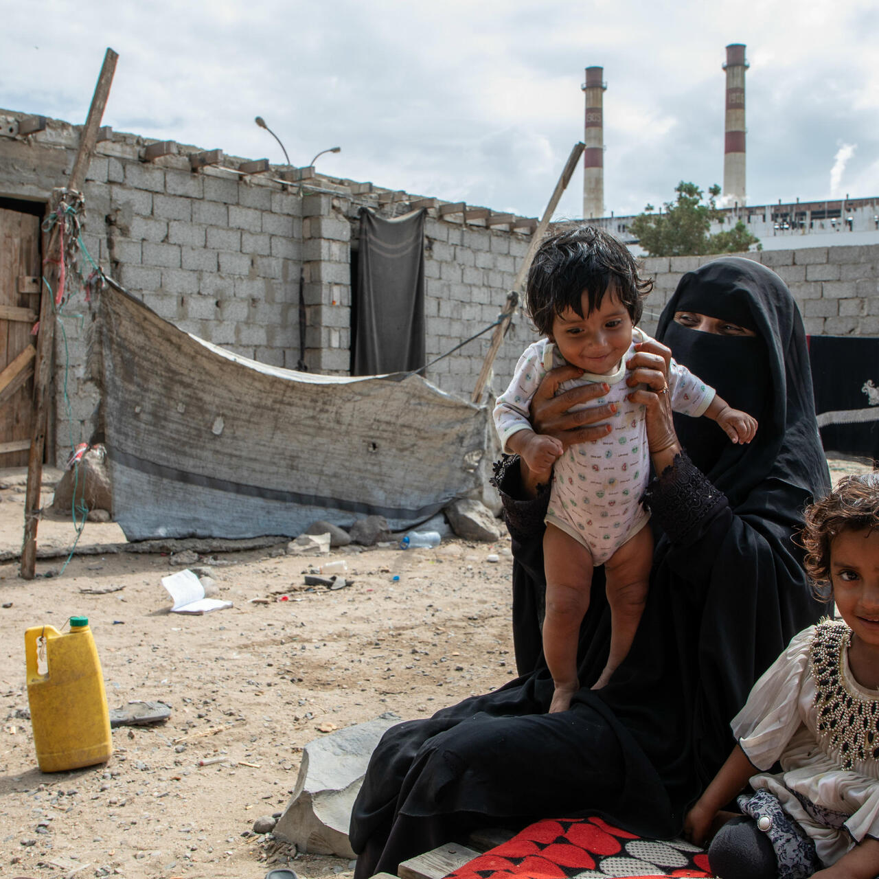 Eine vermummte Frau hält ein Kleinkind (Salman) hoch, neben ihr sitzt ihre andere Tochter. Alle befinden sich in einem ärmlichen Innenhof.