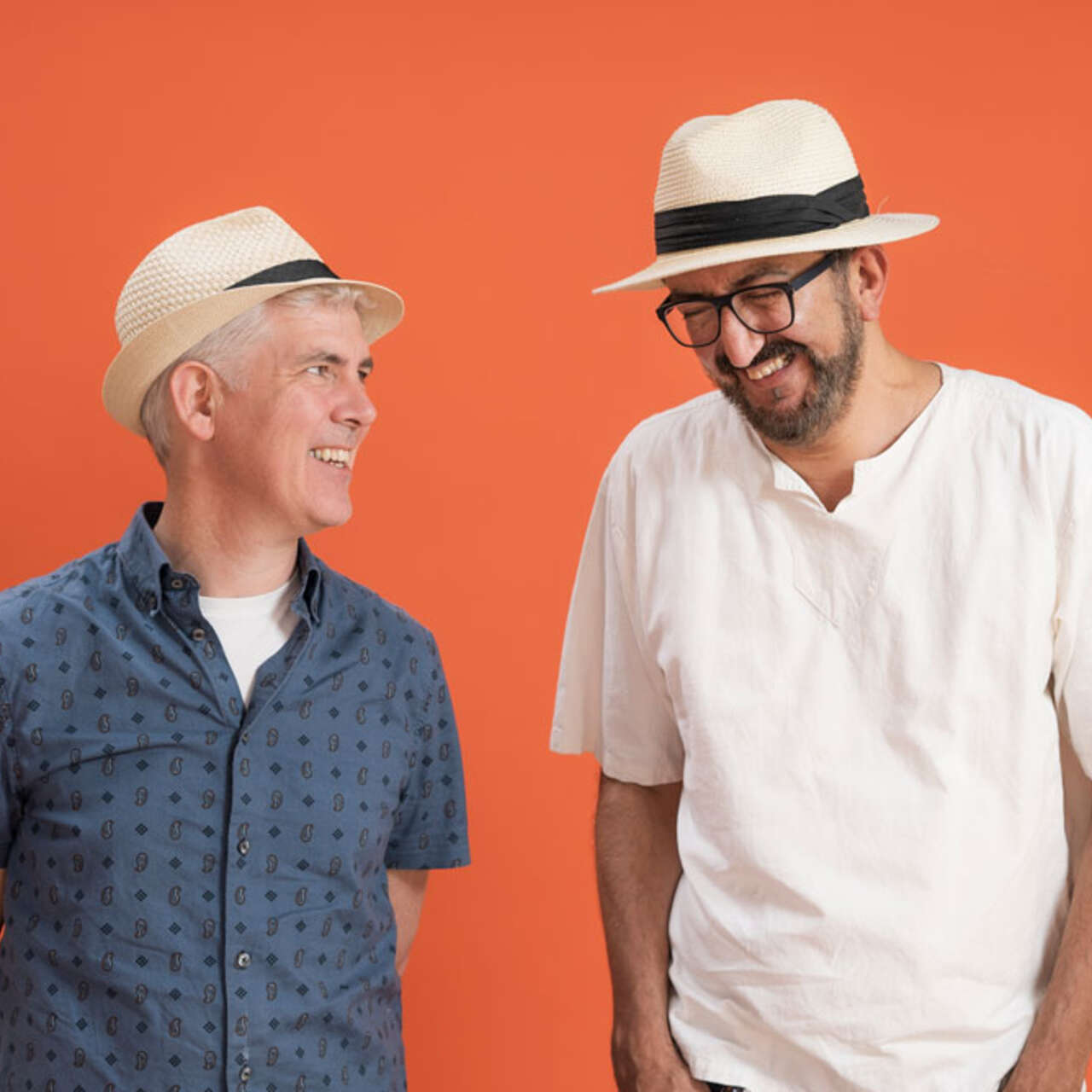 Zwei Männer, die beide Hüte tragen, stehen vor einer orangenen Wand und lachen.
