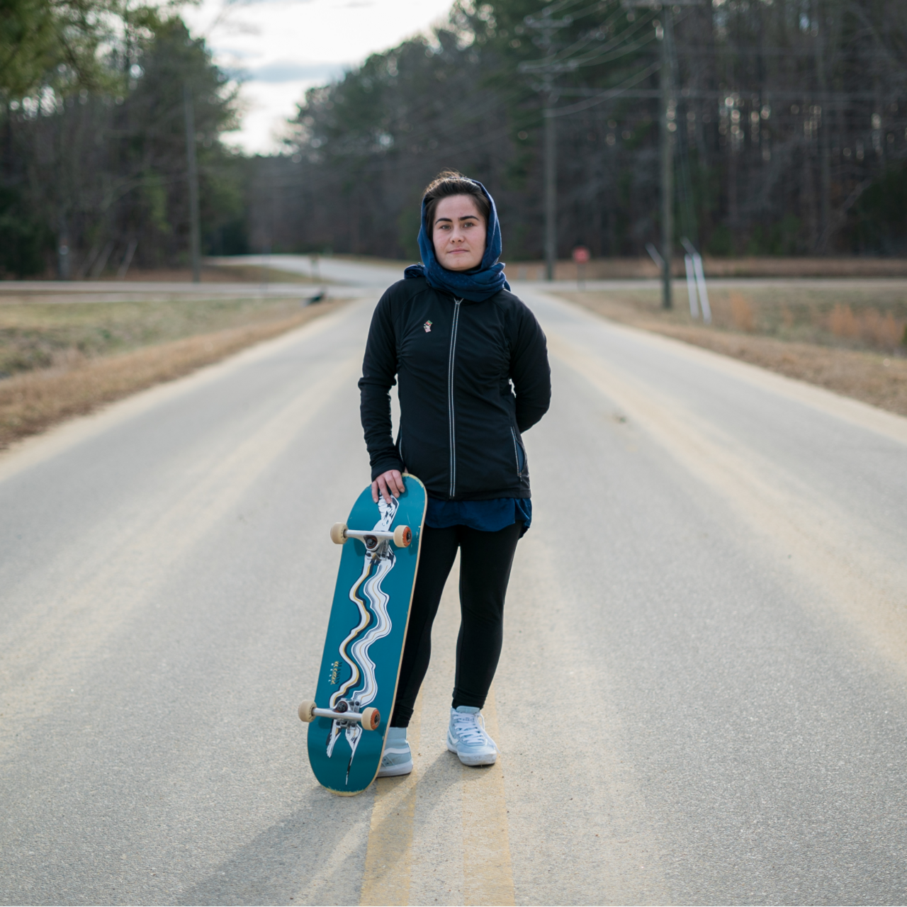 A 22 year old woman stands in the middle of a road holding a skateboard