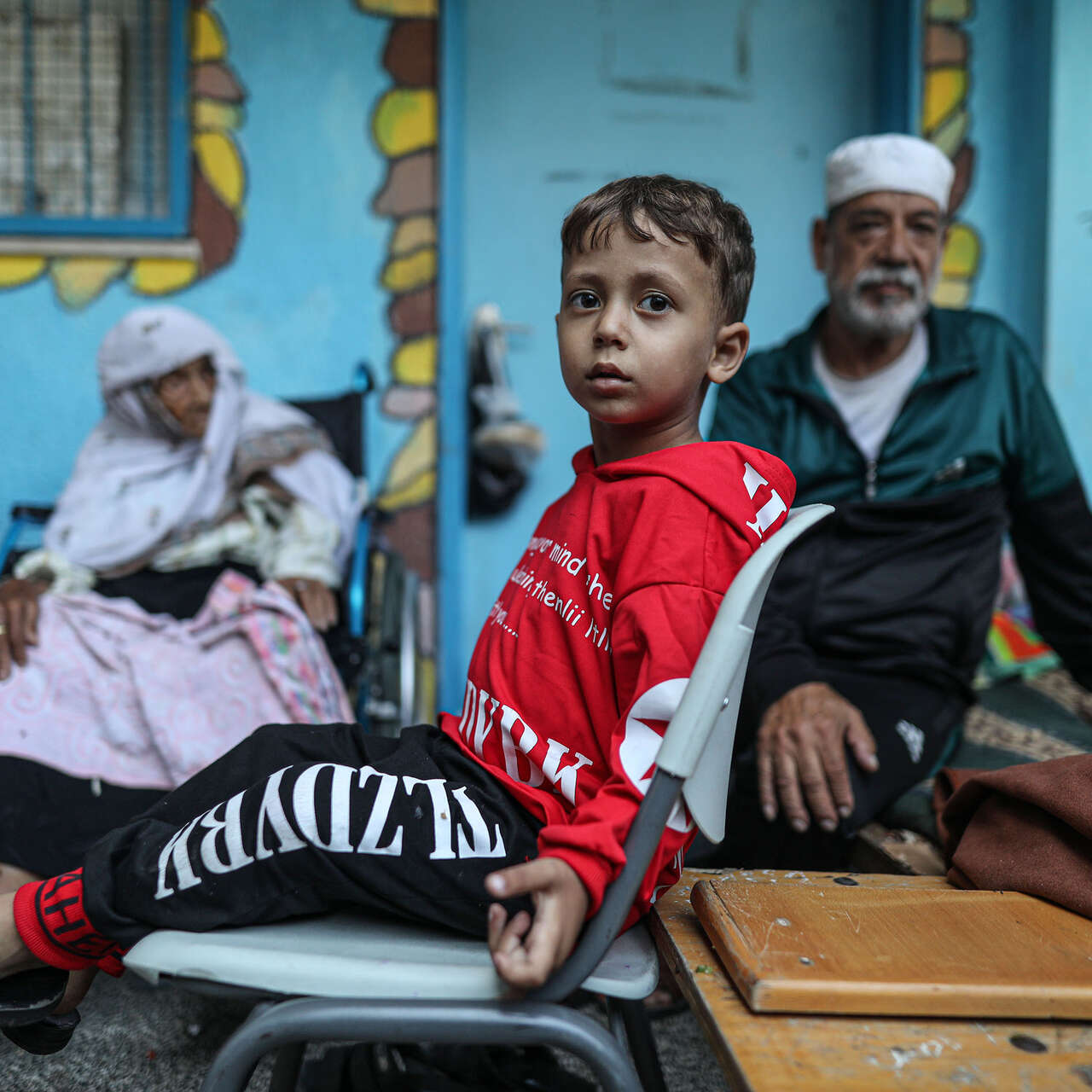 A Palestinian boy sits in a chair at a UNRWA school. An elderly woman and man sit nearby.