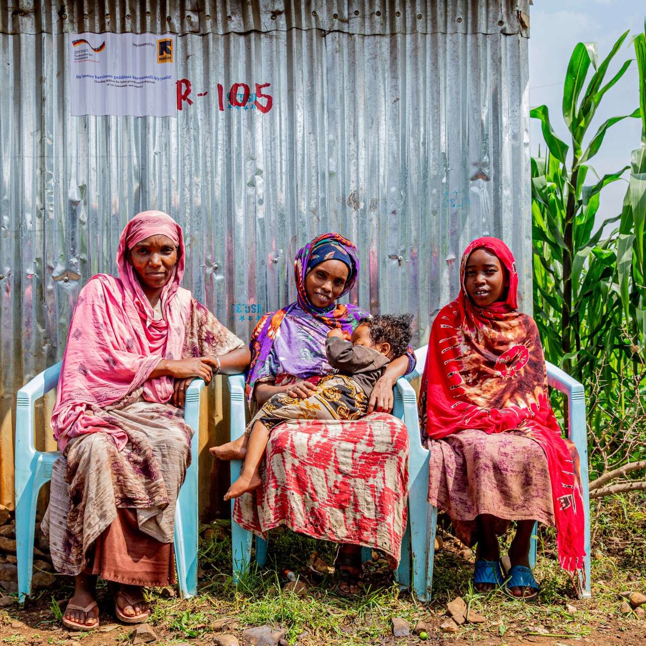 Three women sit in chairs outside and pose for a photo. The woman in the middle holds a young child in her arms.
