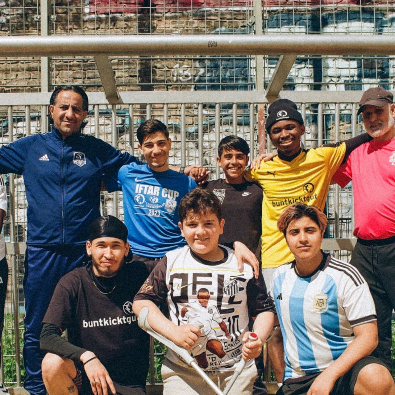 Eine Gruppe von neun Fußballspielern und ihren Trainern steht vor einem Tor auf einem Fußballplatz. Sie lächeln und posieren für das Foto. Rechts unten ist ein Logo mit der Aufschrift 'Football's Coming Home International Rescue Committee' zu sehen.