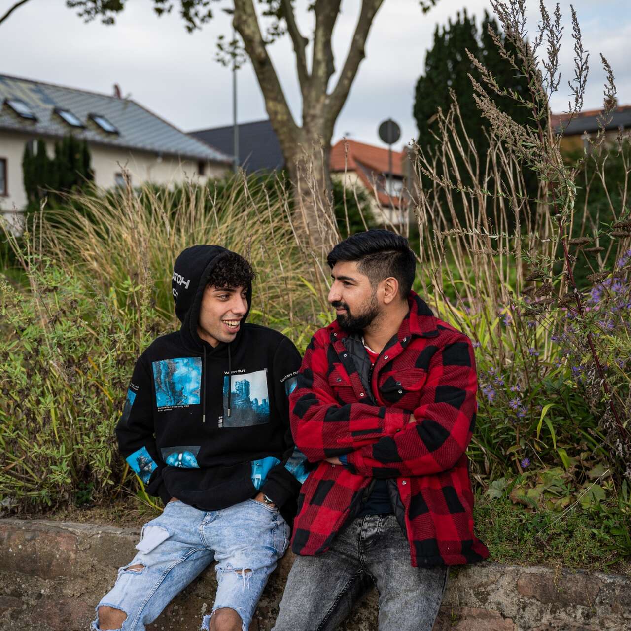 Zwei Menschen sitzen draußen auf einer Bank und unterhalten sich, mit einer grünen Landschaft und Häusern im Hintergrund.