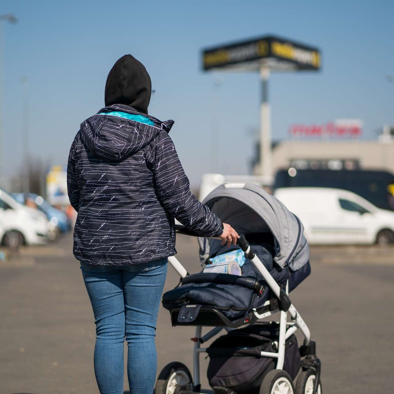 Eine Person, die einen Kinderwagen durch einen Parkplatz schiebt, umgeben von verschiedenen Fahrzeugen und Gebäuden bei klarem Himmel.