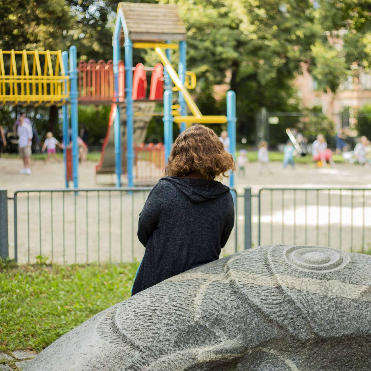 Eine Person sitzt auf einer Bank in einem Park und schaut in Richtung eines Spielplatzes mit bunten Spielgeräten.