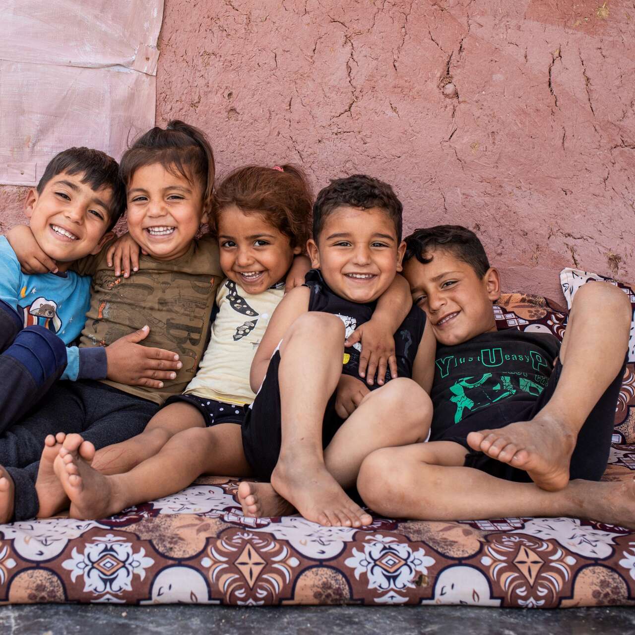 Eine Gruppe von fünf lächelnden Kindern sitzt eng zusammen auf einem gemusterten Sofa und erzeugt eine warme und fröhliche Atmosphäre.