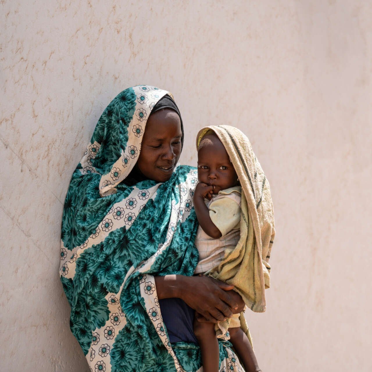 En mamma som flytt från Sudan står lutad mot en vägg och håller sin son i famnen.