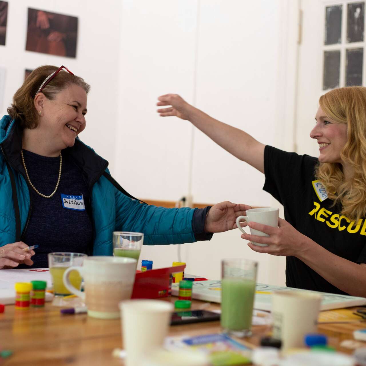 Zwei Frauen sitzen an einem Tisch und lachen zusammen, während sie kreative Materialien vor sich haben.