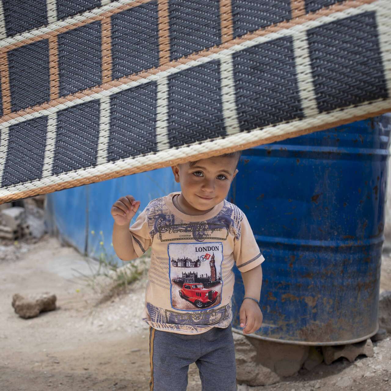 Ein kleines Kind steht in einer provisorischen Unterkunft hinter einer Decke, die als Trennwand dient und hält einen gelben Gegenstand in der Hand.