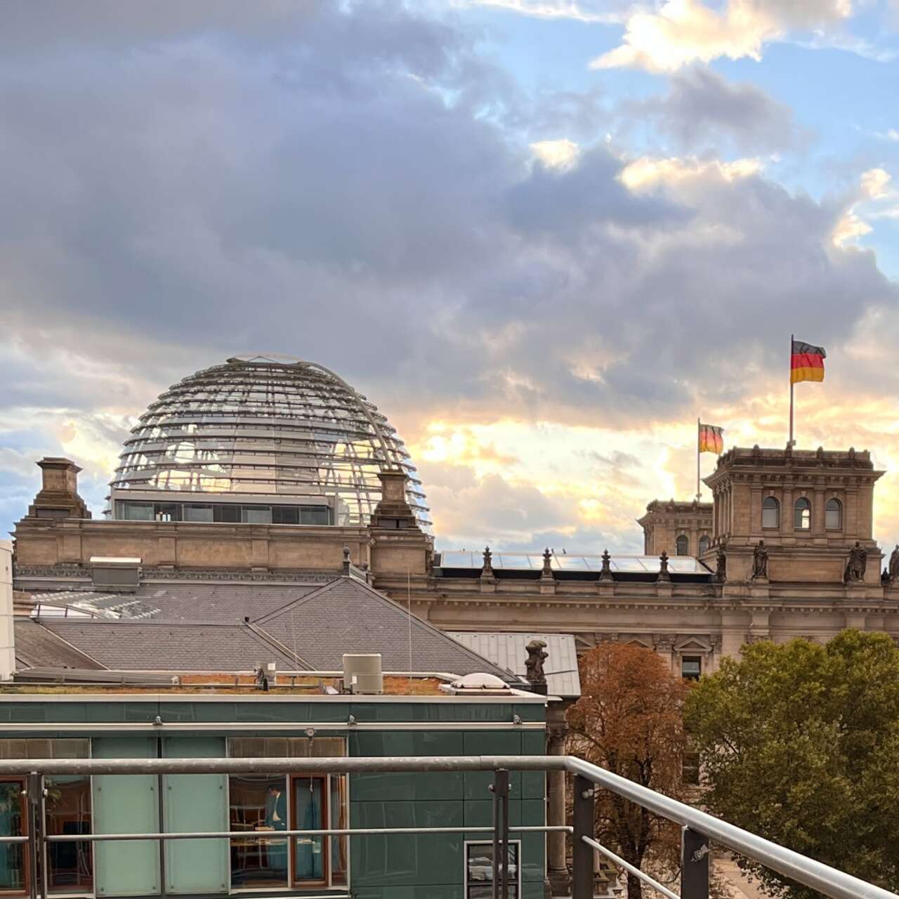 Ein Erwachsener und ein Kind stehen auf einer Terrasse mit Blick auf moderne Gebäude und einen bewölkten Himmel.