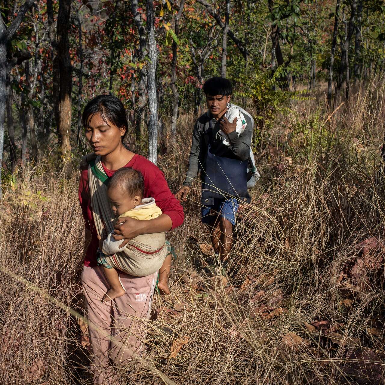 En mamma bär sitt barn i en sele och tar sig fram genom en tätvuxen skog i Myanmar. Kvinnans man går bakom dem.