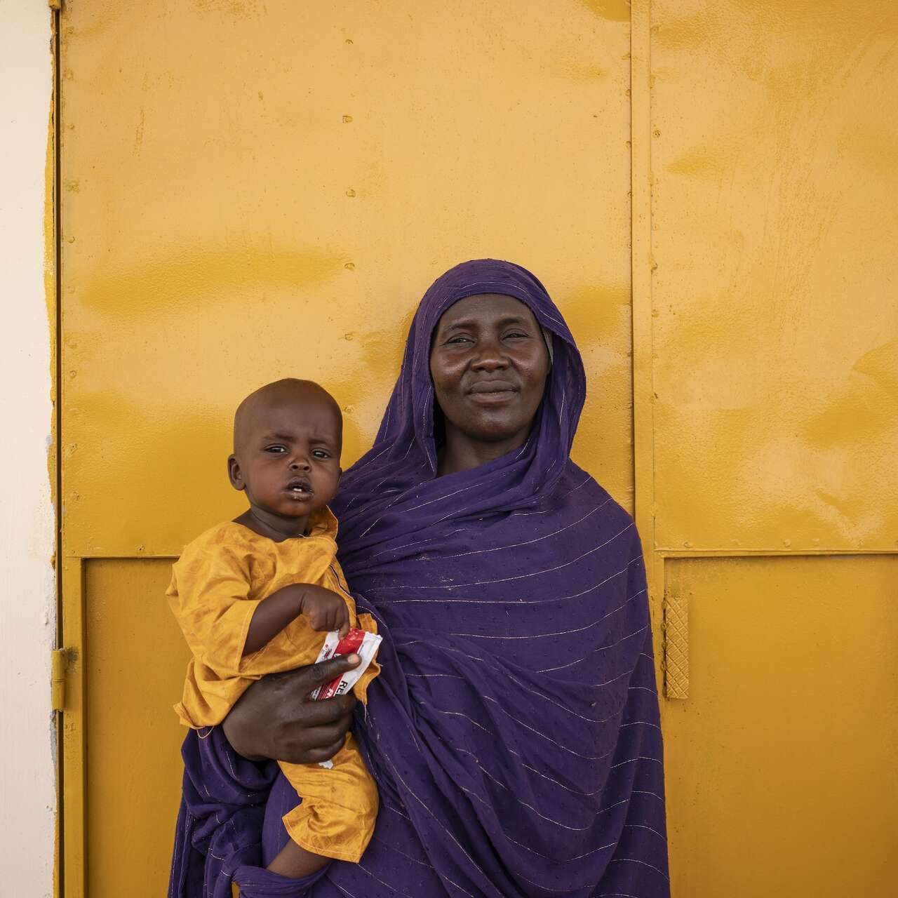 A woman holding her baby against a yellow wall and looking into the camera and smiling