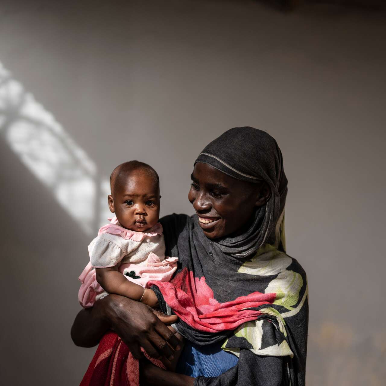 Frau mit Kopftuch hält ein Baby im Arm und lächelt, Lichtstrahl im Hintergrund.