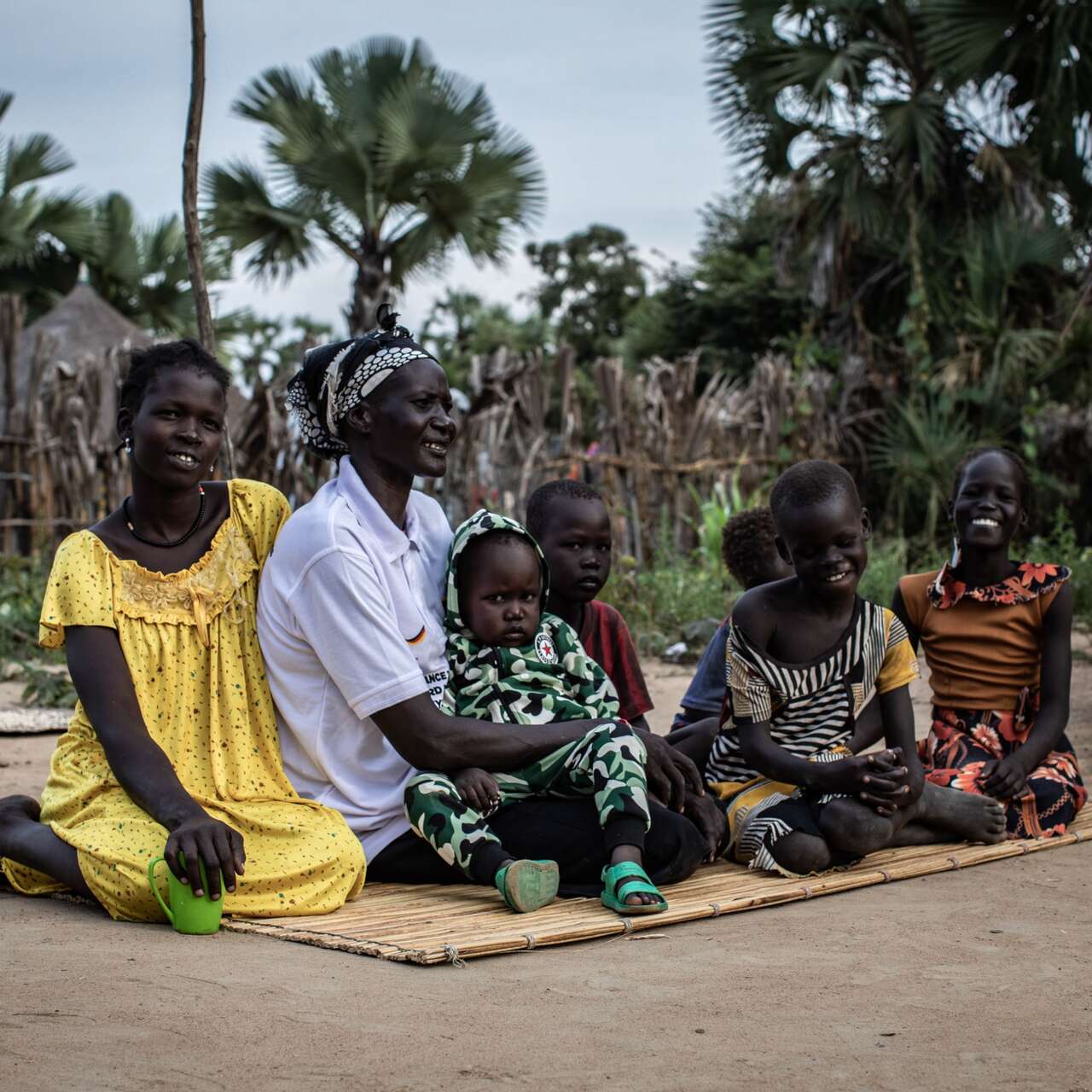 Frau mit Kindern sitzt auf einer Matte vor einem Haus in Südsudan.