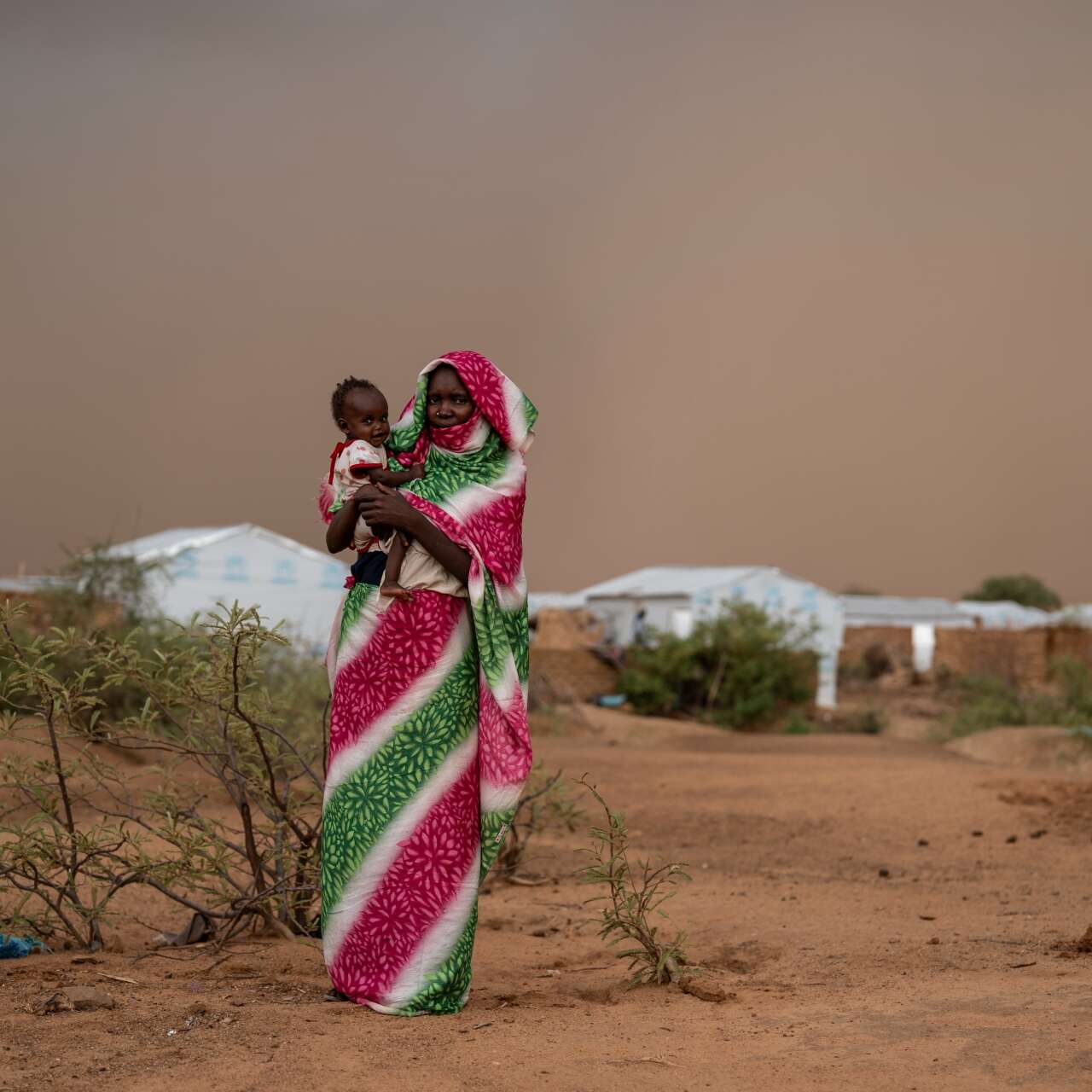 A Sudanese mother holds her young child in her arm. They stand on the outskirts of a camp for displaced people.