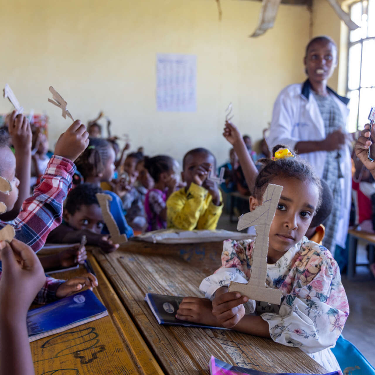 Flera barn sitter i ett klassrum i Etiopien och sträcker upp siffror i luften, i bakgrunden syns deras lärare.