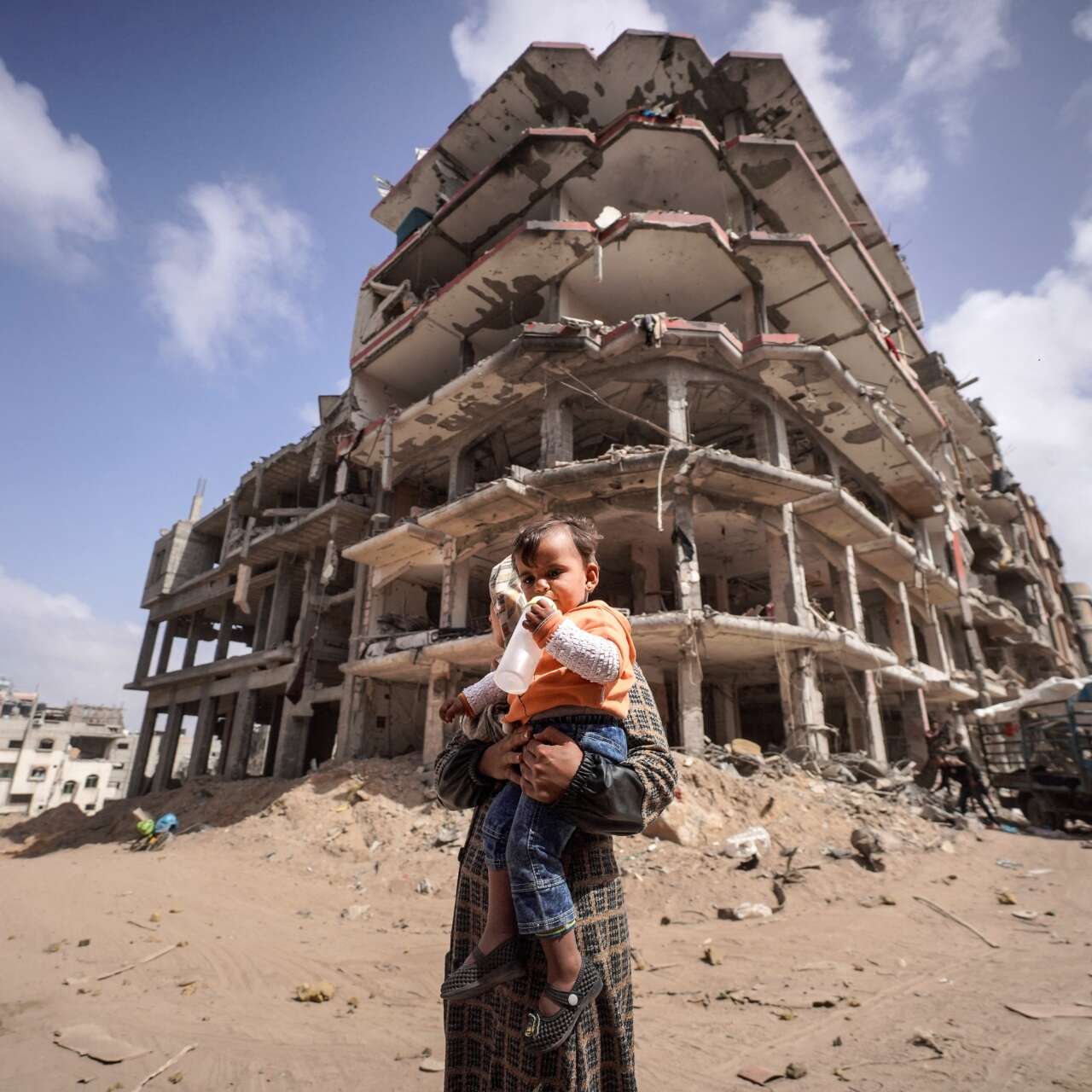 A mother holds a young child in her arms while walking past a destroyed building in Gaza.