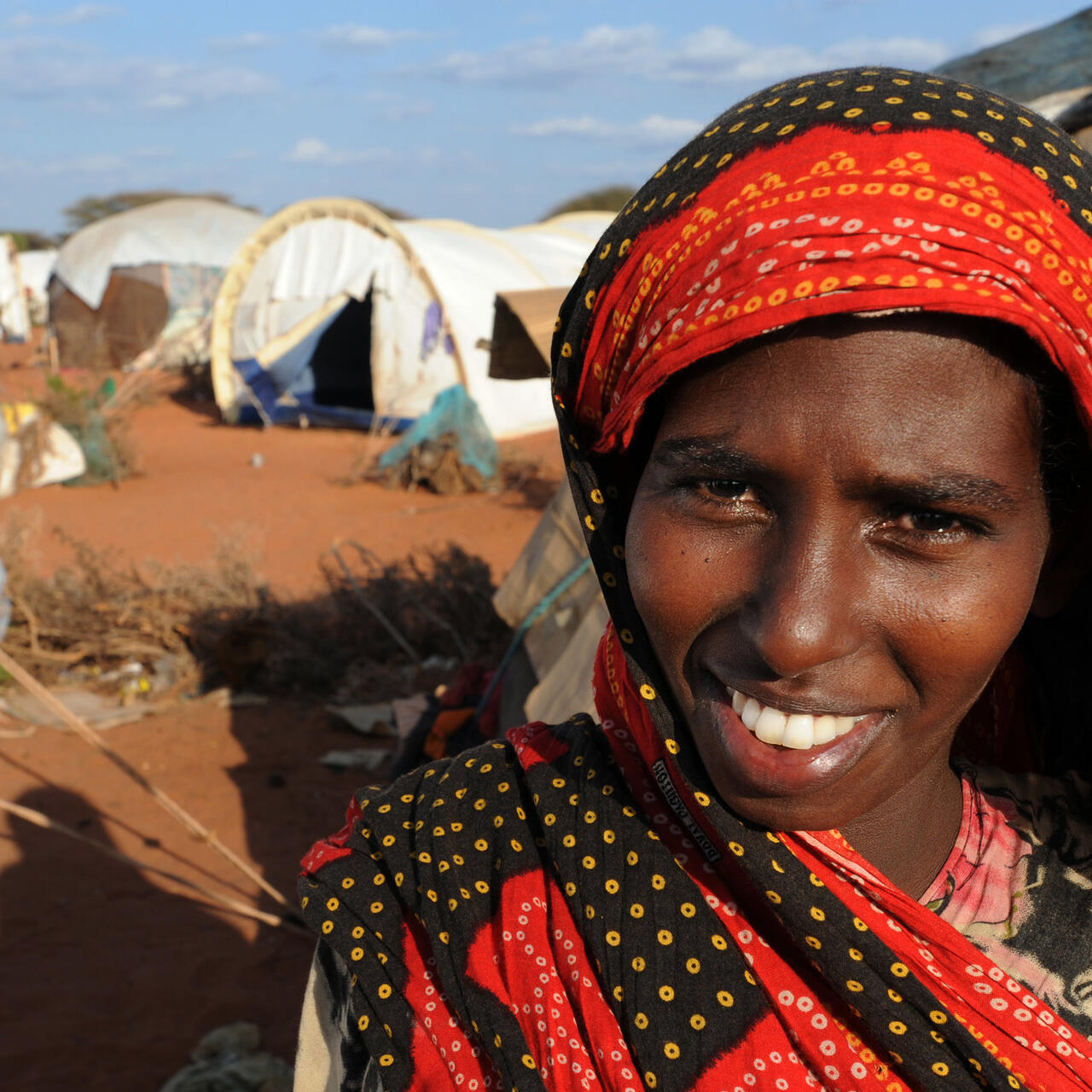 A smiling young woman stands outside a tent in Dadaab refugee camp in Kenya on a sunny day,