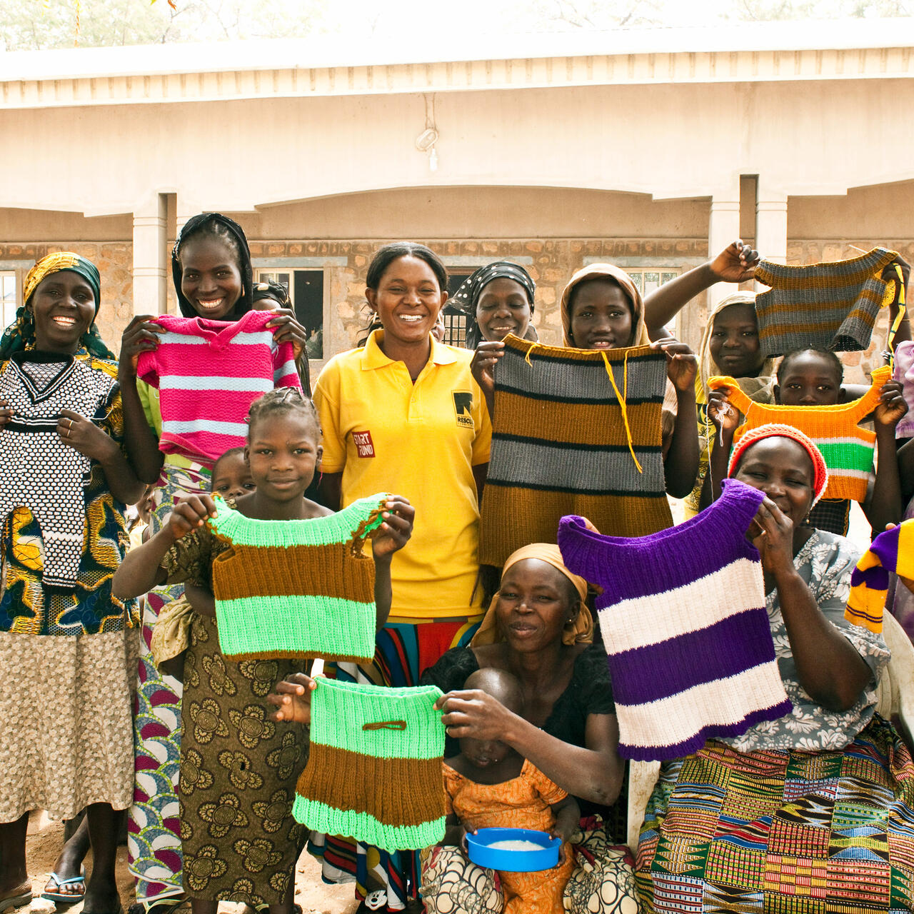 A group of women who are part of an IRC livelihoods program in Nigeria stand outside, lifting hand-knitted tops to show them off.