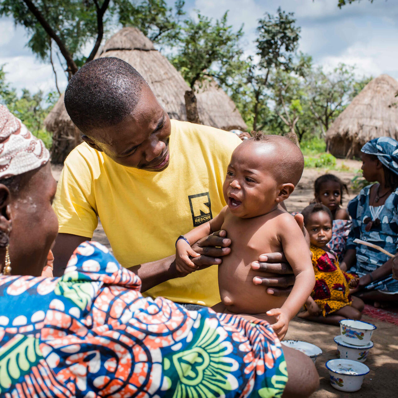 An IRC health worker holds a crying baby, giving him a medical checkup in a village in Central African Republic as his mother looks on smiling.