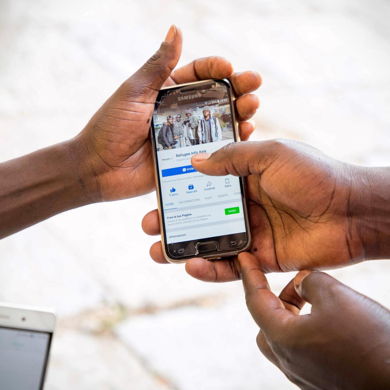 Hands holding a cellphone displaying an IRC Refugee.Info Facebook page on-screen