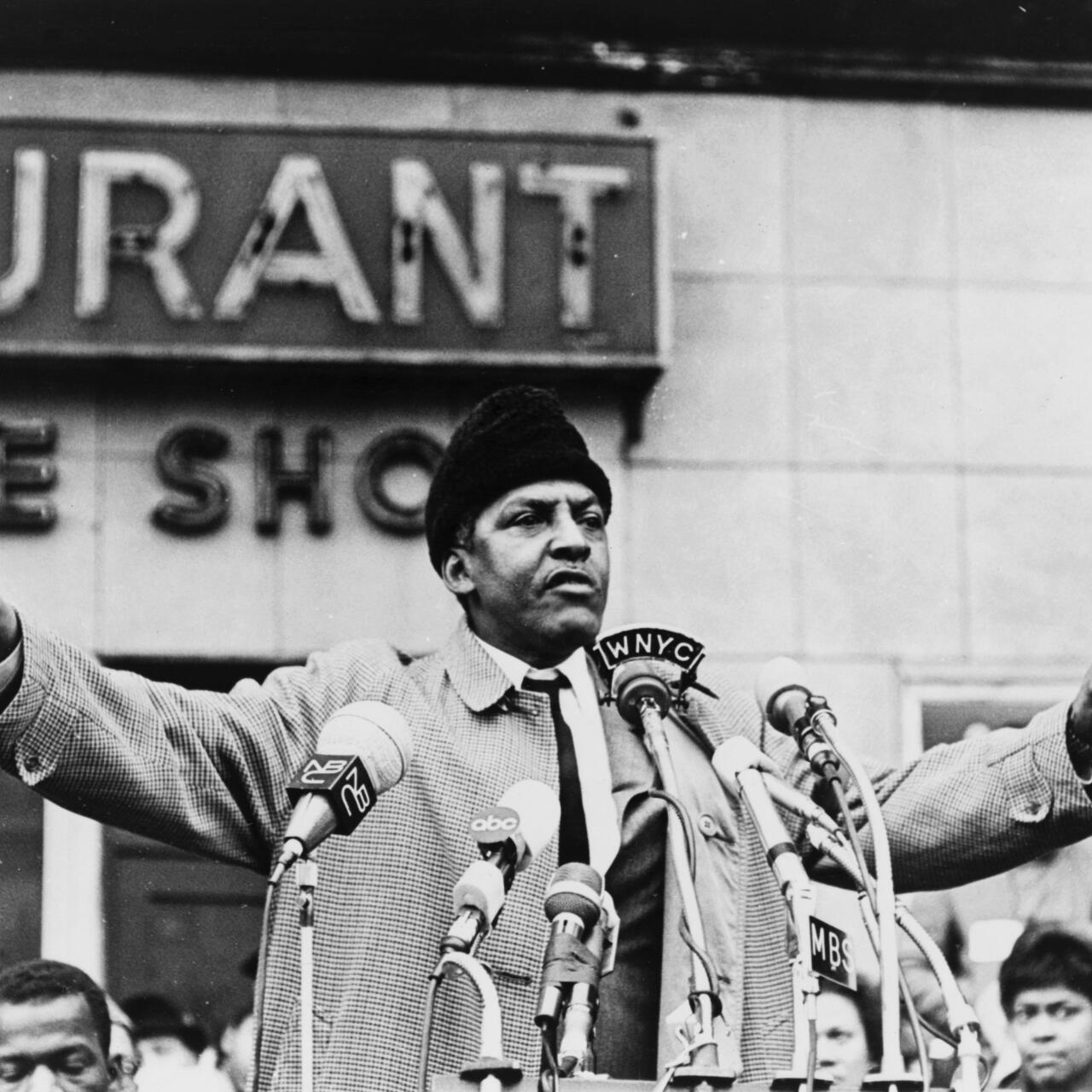 Bayard Rustin stands before microphones at a podium with arms outstretched