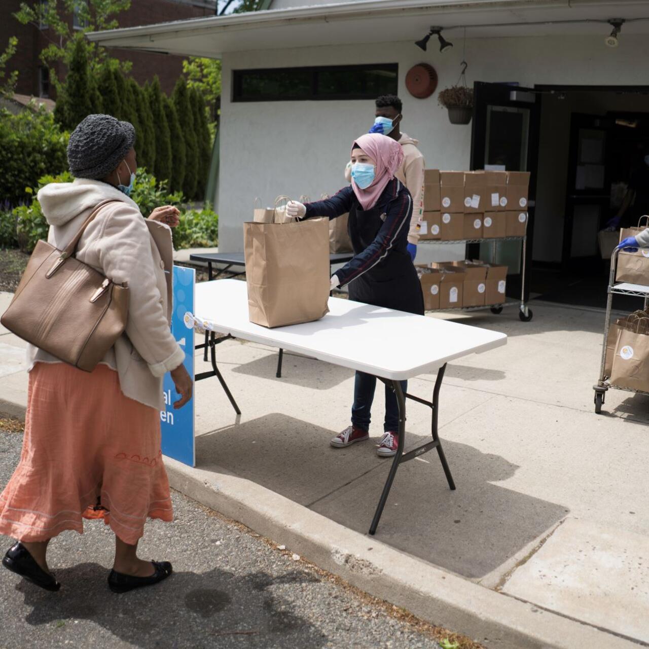 Rania Abou, a nineteen-year-old refugee from Syria, hands out food at her job with WC Kitchen and the IRC in Elizabeth, New Jersey. She is wearing a mask, a pink head scarf, and a black jacket and is handing a paper grocery bag to a woman in a pink skirt.