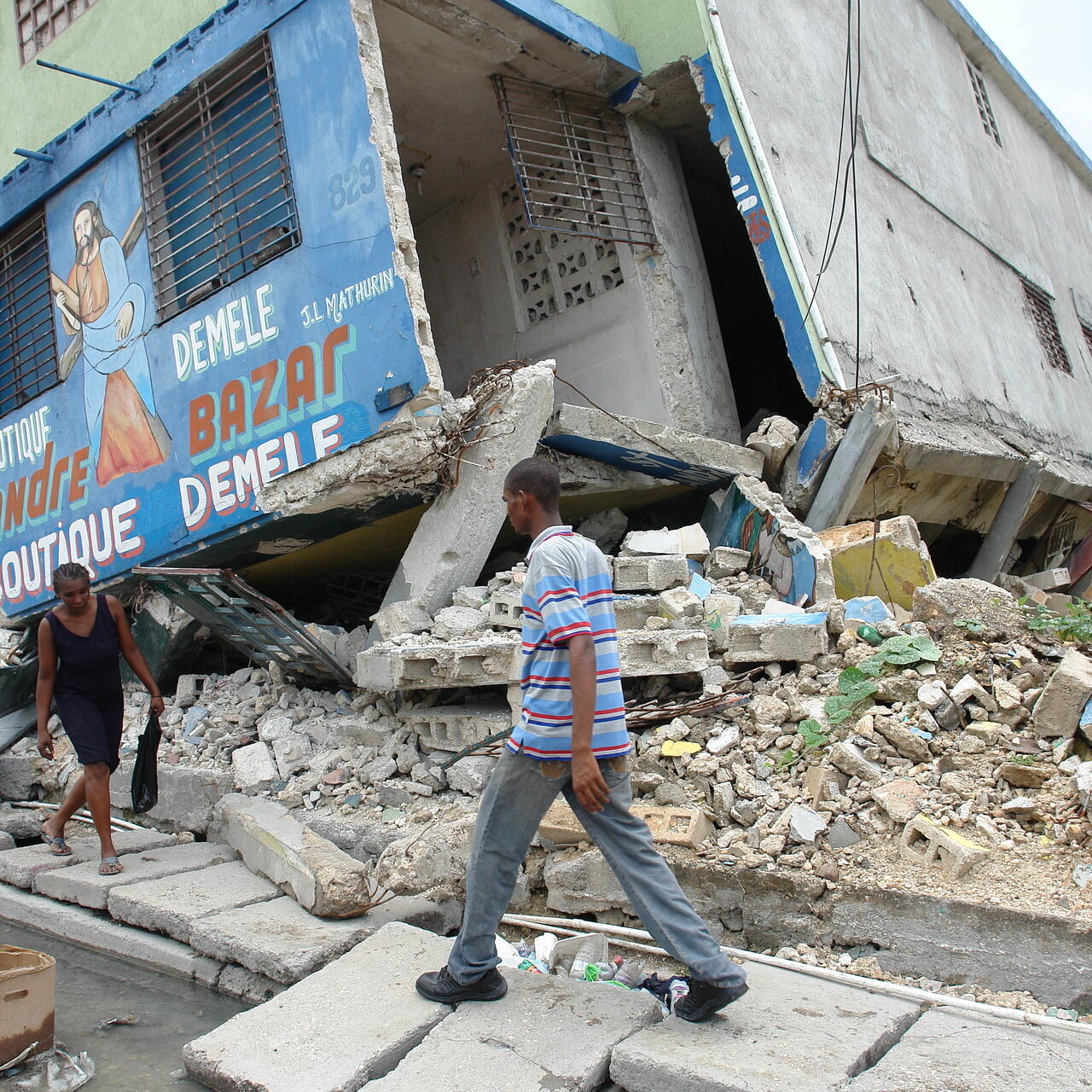 A man and a woman walk carefully on a damaged sidewalk past collapsed buildings in Port-au-Prince, Haiti after the 2010 earthquake.