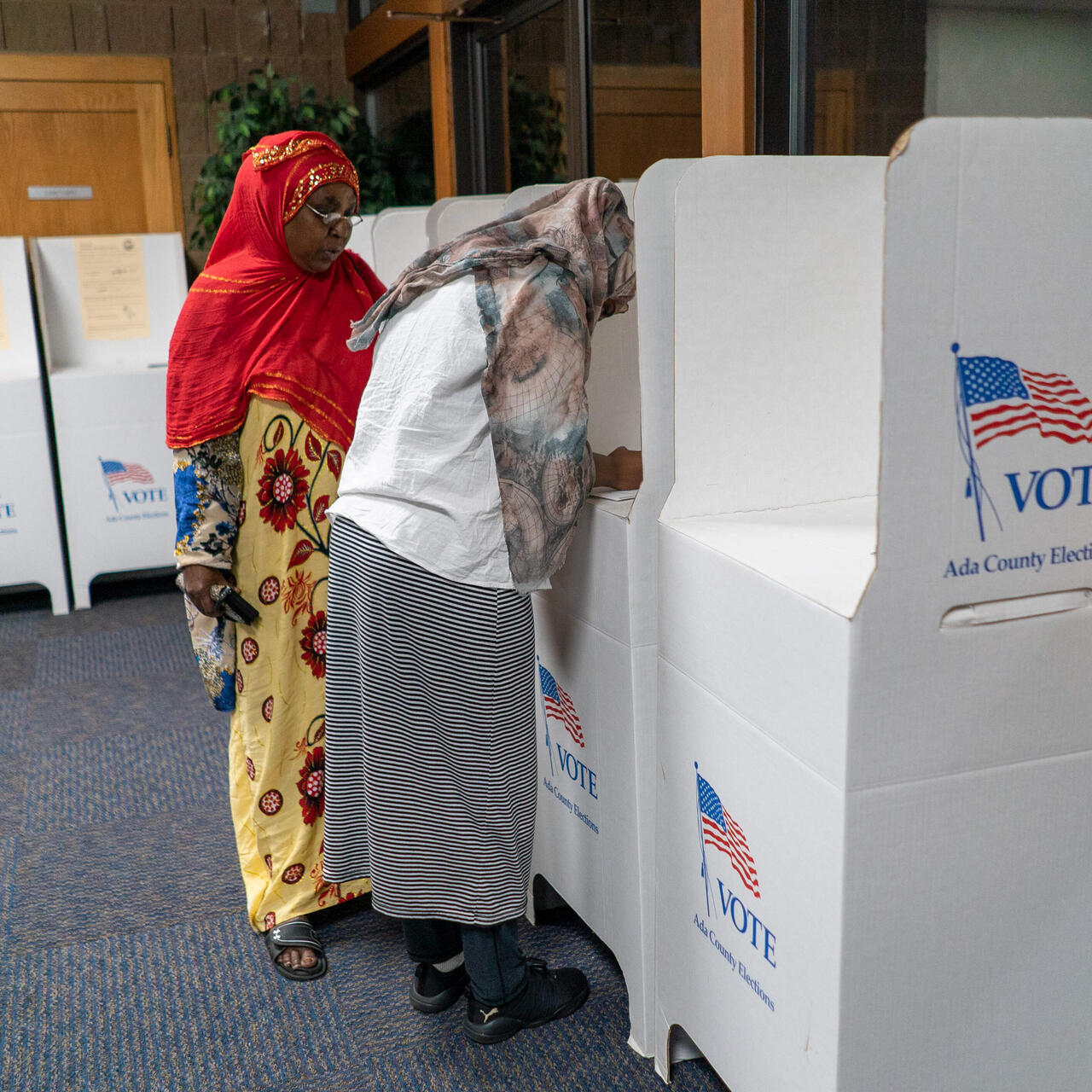 Two woman vote in an election in Boise, Idaho. One is filling out her ballot in a privacy booth while the other stands next to her.