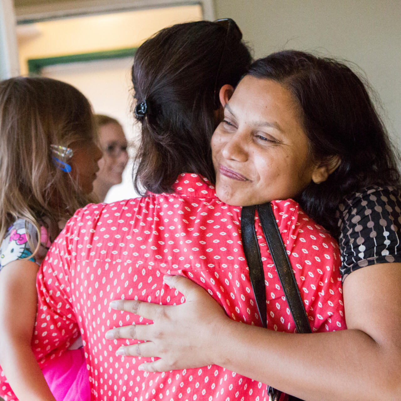 Two women hug while one holds a small child. We see one of their faces, she is smiling.